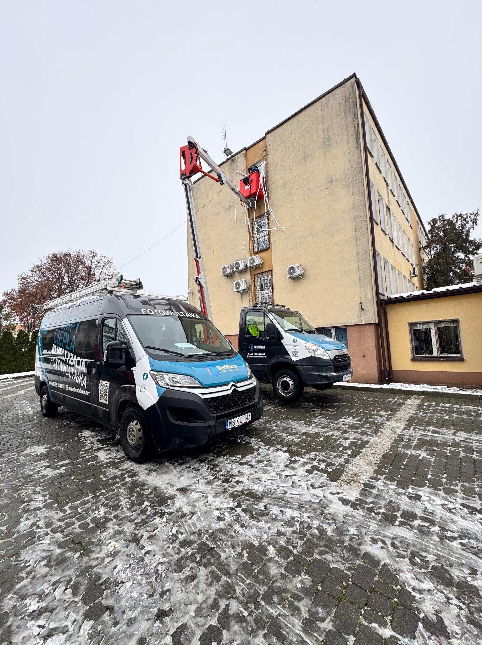 Montaż klimatyzacji na budynku z podnośnika koszowego. Dwa samochody firmowe z logo i hasłami reklamowymi na pierwszym planie. Zimowa sceneria.