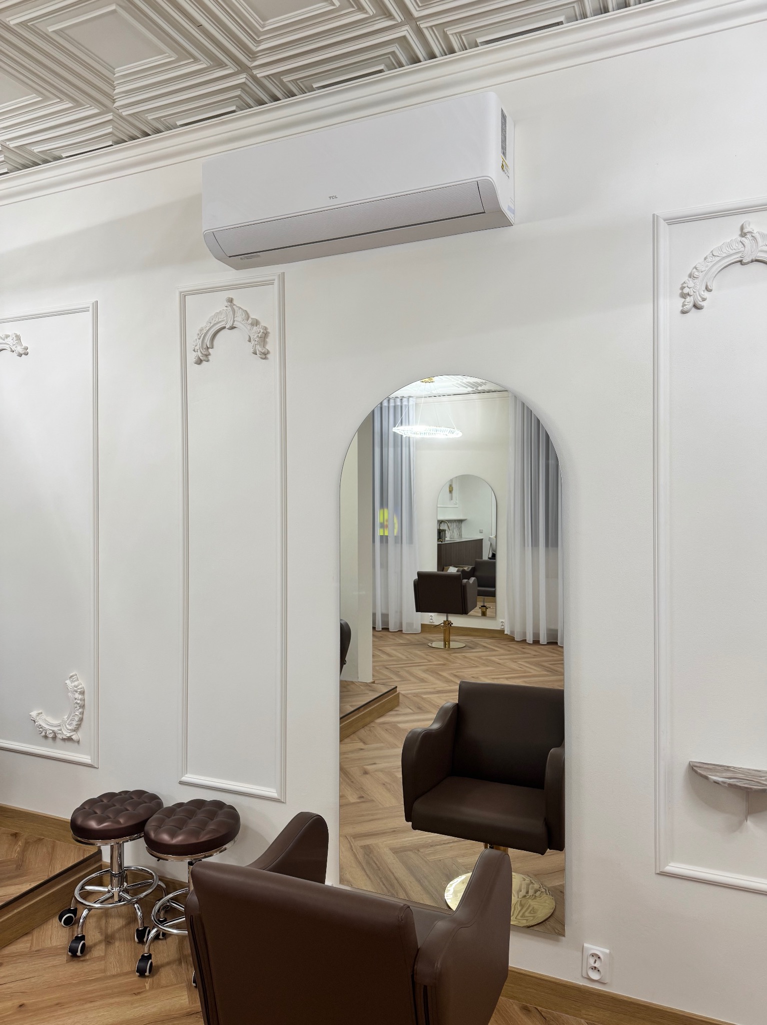 Białe wnętrze salonu z klimatyzatorem TCL na ścianie, widoczne krzesła fryzjerskie i lustro w kształcie łuku. Dekoracyjne sztukaterie na ścianach i kasetonowy sufit.