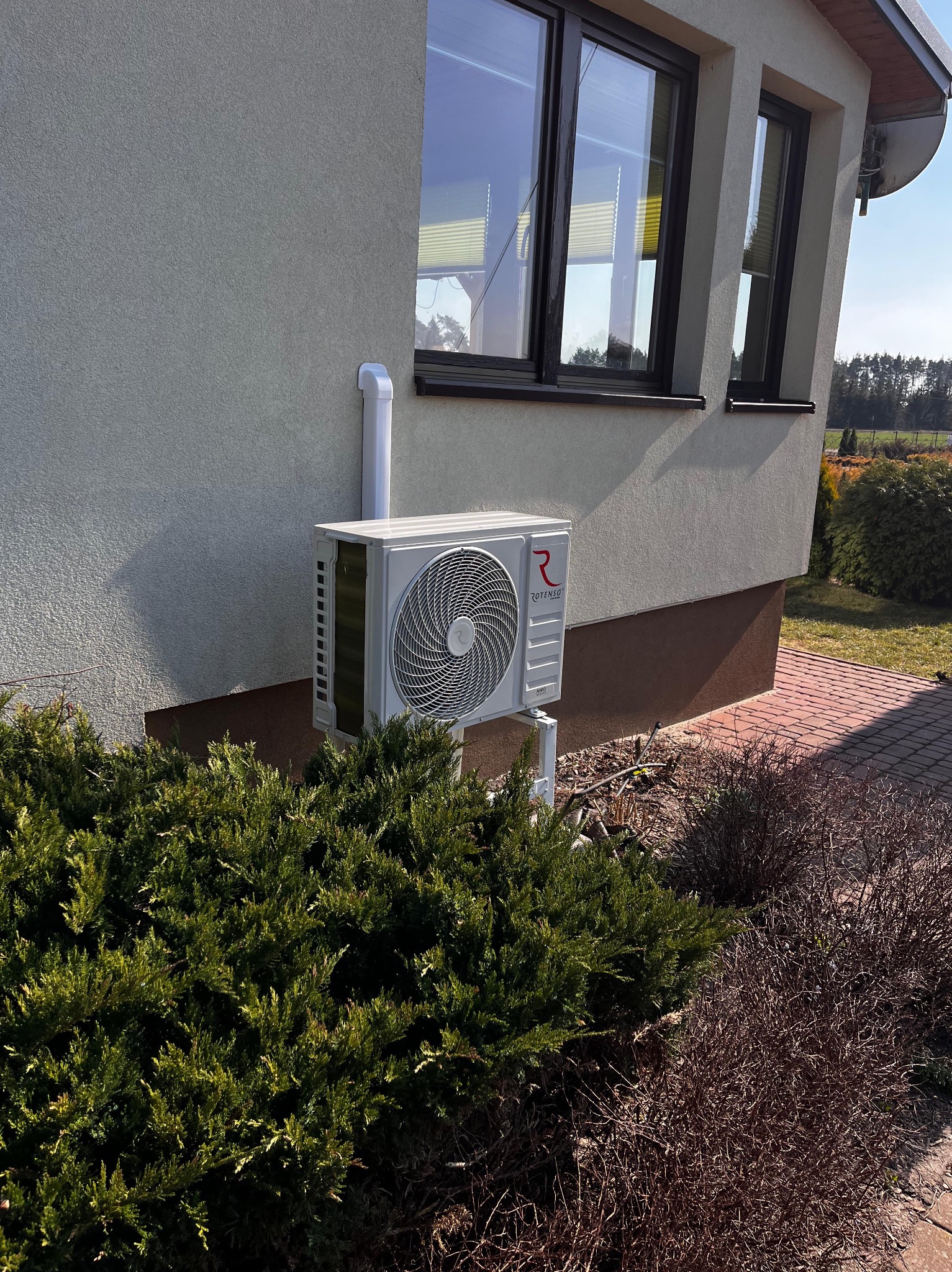 Biały klimatyzator Rotenso zamontowany na elewacji domu, z widoczną instalacją i otoczeniem zieleni. Ujęcie z poziomu gruntu.