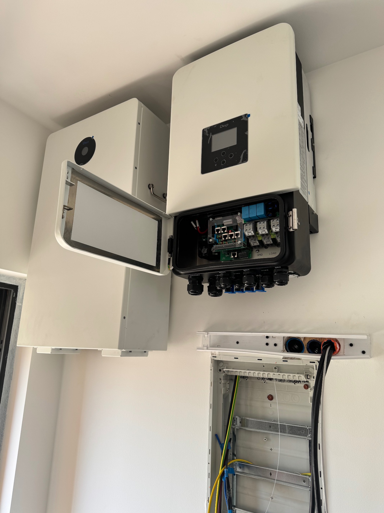 Biały inwerter fotowoltaiczny Deye z otwartą obudową, widoczne okablowanie i podłączenia, obok akumulator energii i skrzynka elektryczna na białej ścianie.