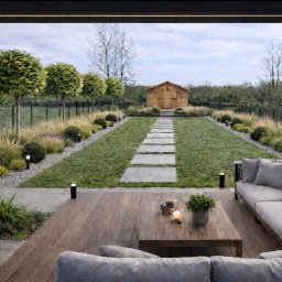 Witold Bączek - Nowoczesny ogród z drewnianym tarasem i szarą sofą. Ścieżka z płyt prowadzi do drewnianego domku. Oświetlenie ogrodowe podkreśla roślinność i architekturę.