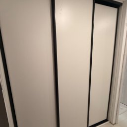 sebastian schönwetter - Zamknięta, trzydrzwiowa szafa przesuwna z białymi frontami i czarnymi obramowaniami, widoczna z perspektywy pod lekkim kątem, z fragmentem podłogi w kadrze.