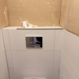 sebastian schönwetter - Świeżo wykafelkowana ściana w łazience z zamontowanym stelażem WC i przyciskiem spłukującym. Nad płytkami widoczna surowa ściana przygotowana do malowania.