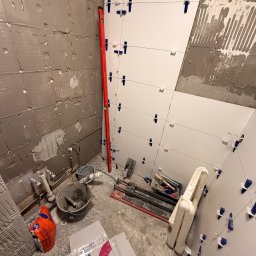 sebastian schönwetter - Układanie białych płytek w łazience, widoczne narzędzia glazurnicze, poziomica i fragment ściany przygotowanej pod płytki. Etap prac wykończeniowych.