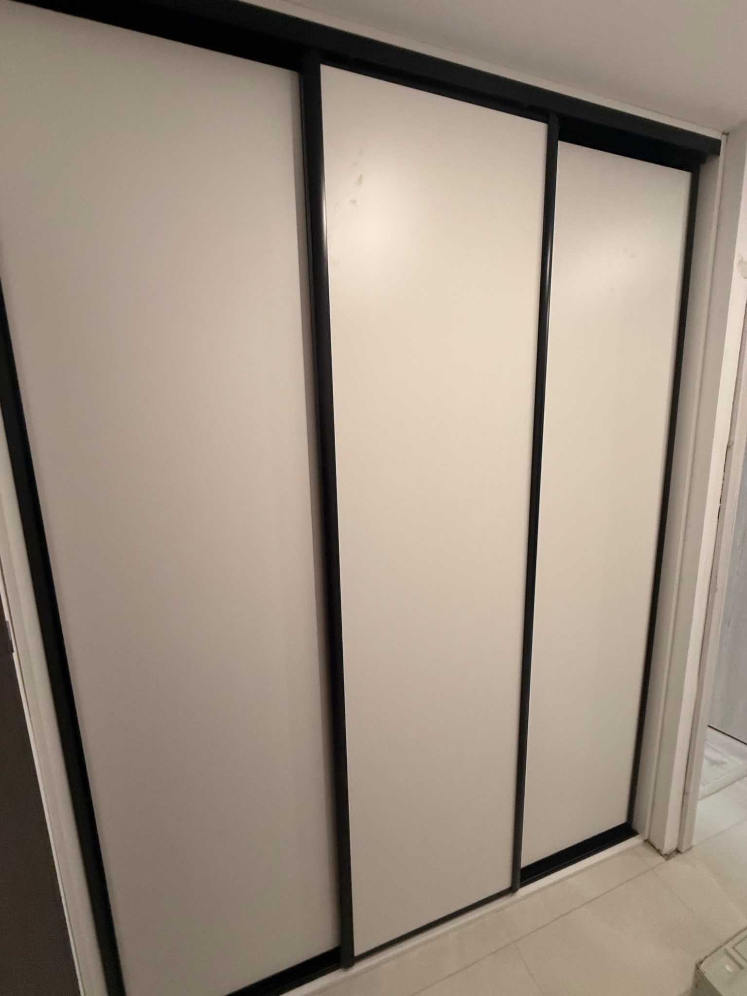 Zamknięta, trzydrzwiowa szafa przesuwna z białymi frontami i czarnymi obramowaniami, widoczna z perspektywy pod lekkim kątem, z fragmentem podłogi w kadrze.