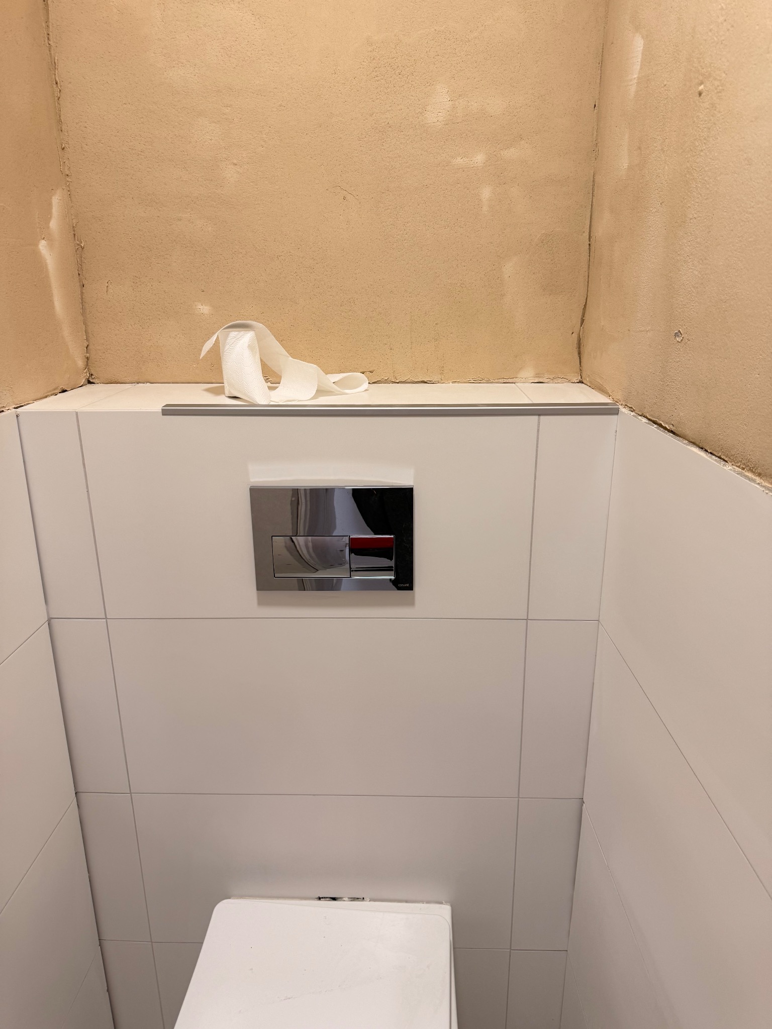 Świeżo wykafelkowana ściana w łazience z zamontowanym stelażem WC i przyciskiem spłukującym. Nad płytkami widoczna surowa ściana przygotowana do malowania.