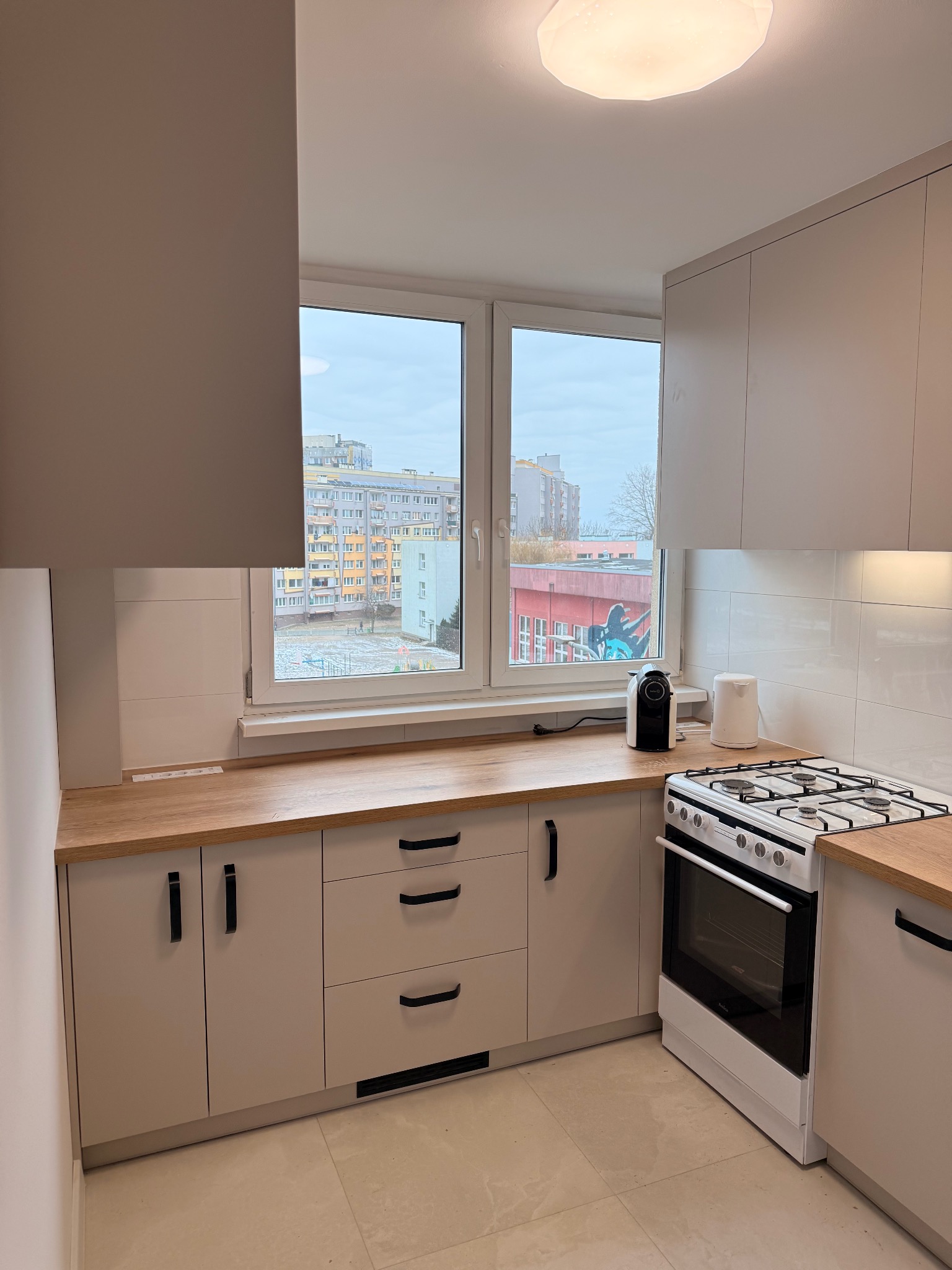 Jasna kuchnia z drewnianym blatem, widokiem na blok zza okna i nowoczesnym oświetleniem sufitowym. Minimalistyczny design, sprzęt AGD na blacie.