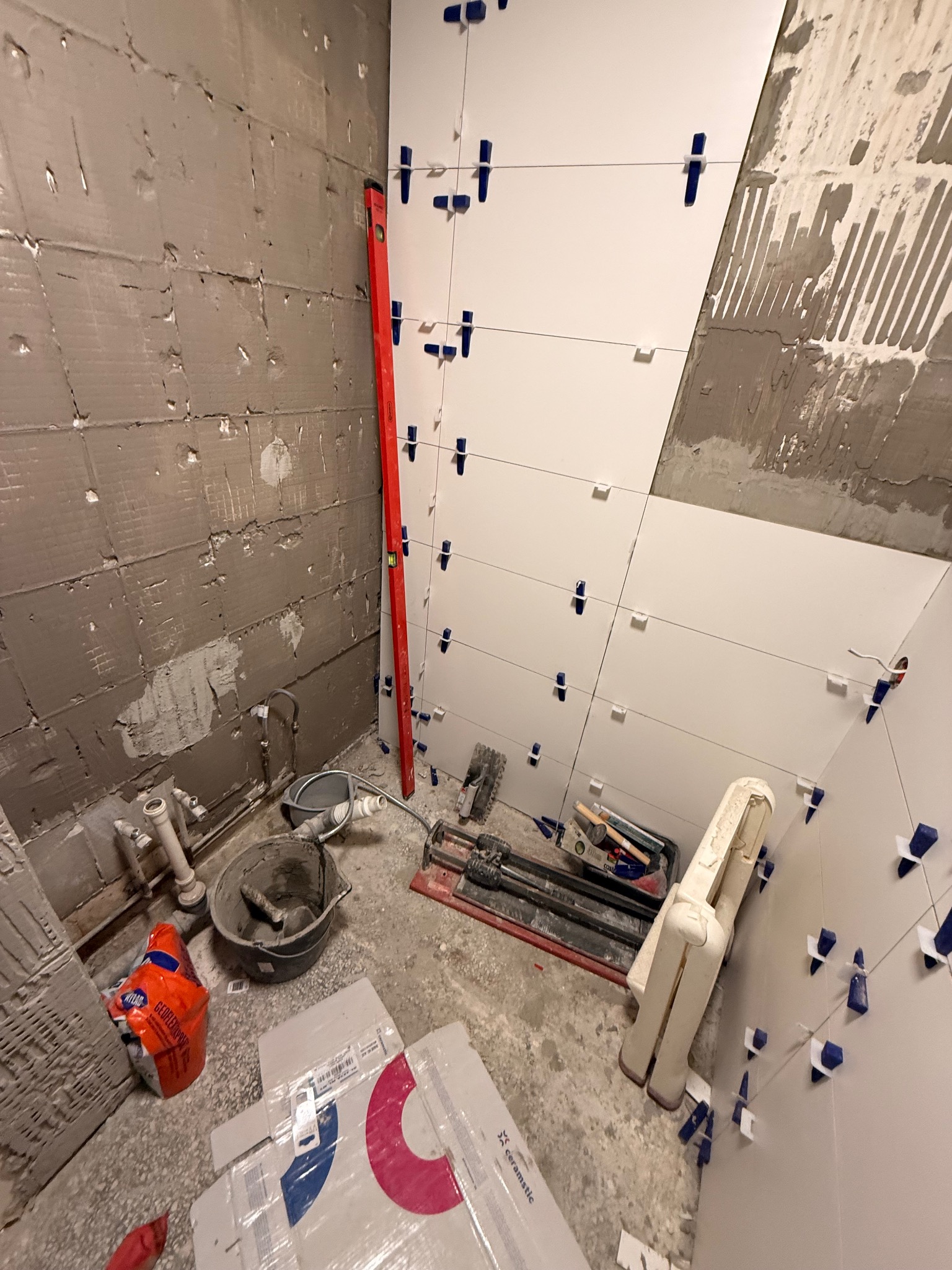 Układanie białych płytek w łazience, widoczne narzędzia glazurnicze, poziomica i fragment ściany przygotowanej pod płytki. Etap prac wykończeniowych.