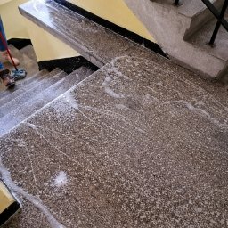 Robota na Glanc - Czyszczenie schodów z lastryko z użyciem mopa i detergentu. Widoczne ślady piany i wilgoci na stopniach. Fragment osoby z mopem w lewym górnym rogu kadru.