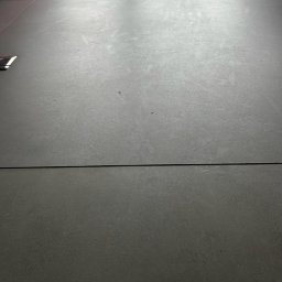 Robota na Glanc - Szara, matowa podłoga z płytek, widoczne fugi i delikatna tekstura. W tle fragmenty mebli i dekoracji, sugerujące minimalistyczne wnętrze. Stonowana kolorystyka.