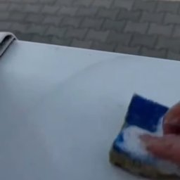 Robota na Glanc - Czyszczenie powierzchni gąbką z detergentem. Widoczna dłoń w trakcie pracy, fragment kostki brukowej w tle. Ujęcie z bliska, skupione na procesie usuwania zabrudzeń.