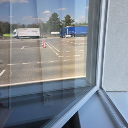 Robota na Glanc - Widok z okna na parking z ciężarówkami i stożkami. Biała rama okienna z widocznym gniazdkiem elektrycznym. Częściowe odbicie wnętrza w szybie.
