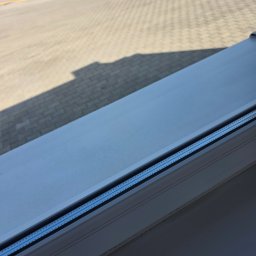 Robota na Glanc - Widok z okna na kostkę brukową i słupki drogowe w słoneczny dzień. Okno z widocznymi detalami ramy i uszczelki. Cień na kostce brukowej.