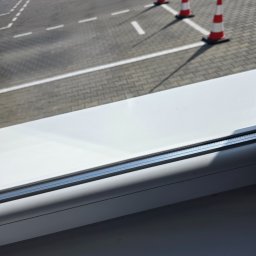 Robota na Glanc - Widok z okna na plac manewrowy z kostki brukowej, wyznaczonymi liniami i ustawionymi pachołkami drogowymi. Perspektywa z góry, zza ramy okiennej.