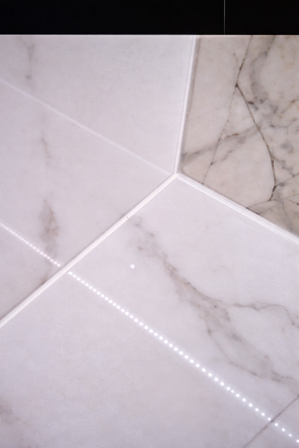Białe i beżowe marmurowe płytki ułożone pod kątem, z widoczną listwą LED wzdłuż spoin. Detal wykończenia podłogi lub ściany.