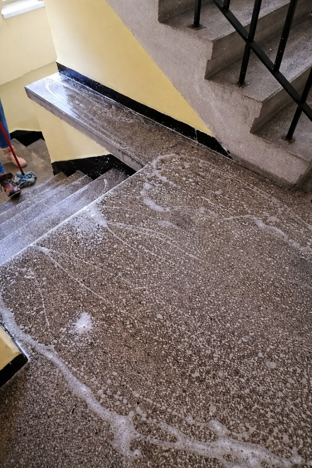 Czyszczenie schodów z lastryko z użyciem mopa i detergentu. Widoczne ślady piany i wilgoci na stopniach. Fragment osoby z mopem w lewym górnym rogu kadru.