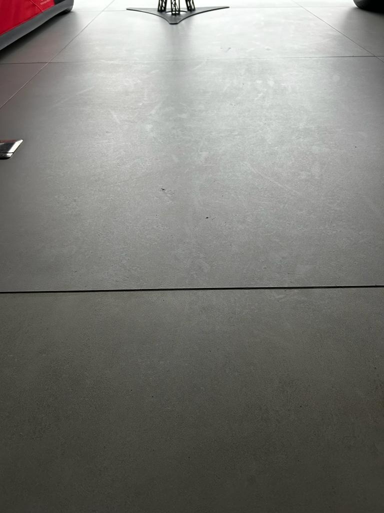 Szara, matowa podłoga z płytek, widoczne fugi i delikatna tekstura. W tle fragmenty mebli i dekoracji, sugerujące minimalistyczne wnętrze. Stonowana kolorystyka.