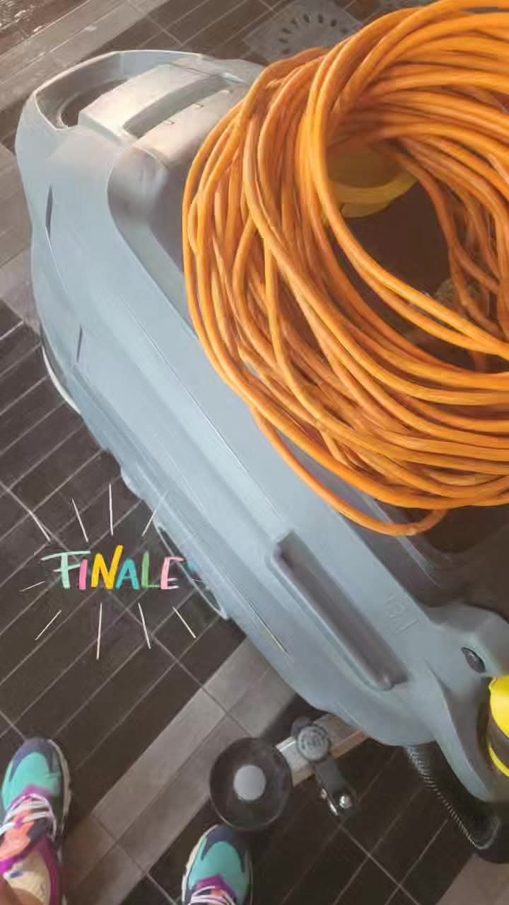 Profesjonalna maszyna czyszcząca z pomarańczowym kablem na podłodze w Chorzowie. Widoczne buty osoby obsługującej urządzenie. Sprzęt do sprzątania.