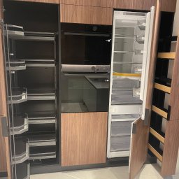 FURNIGO - Nowoczesna kuchnia z drewnianymi frontami: zabudowa z piekarnikiem, lodówką i wysuwanymi półkami cargo. Minimalistyczny design, jasna podłoga.