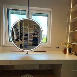 FURNIGO - Beżowa garderoba na wymiar z oświetleniem LED, widoczna w okrągłym lustrze. Na blacie dekoracje i gniazdka elektryczne. W tle widok z okna na zieleń.