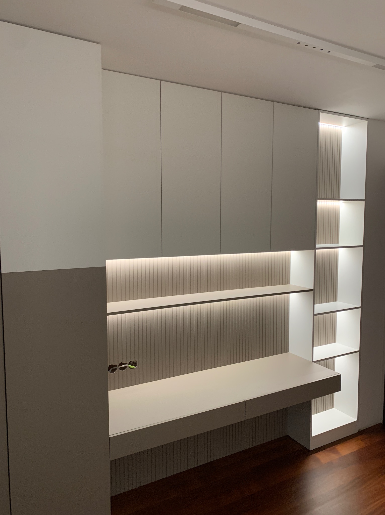 Biała zabudowa meblowa na wymiar z biurkiem i półkami, oświetlenie LED, pionowe żłobienia na ścianie, widok na podłogę. Realizacja z Warszawy.