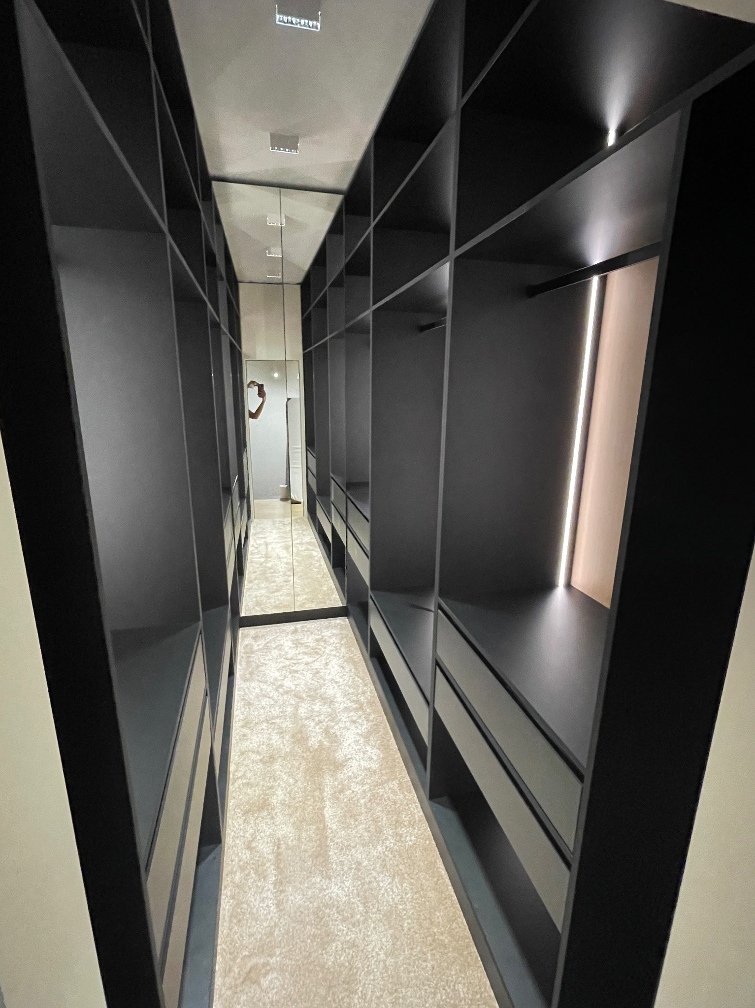 Nowoczesna garderoba na wymiar z czarnymi półkami i szufladami, oświetleniem LED i lustrem na końcu, tworząca elegancką i funkcjonalną przestrzeń do przechowywania.