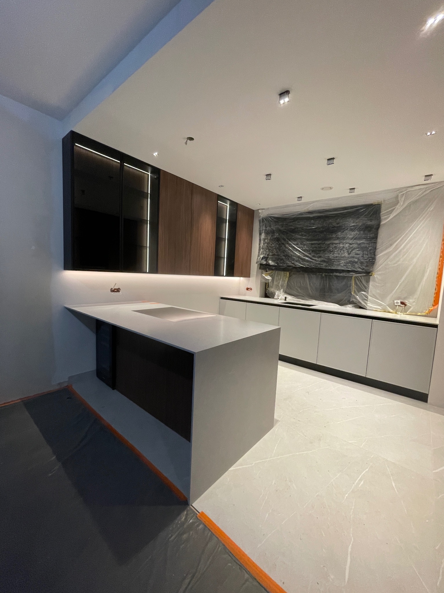 Nowoczesna kuchnia z wyspą, fronty szafek w odcieniach szarości i drewna, oświetlenie LED. Okno zabezpieczone folią ochronną podczas prac wykończeniowych.