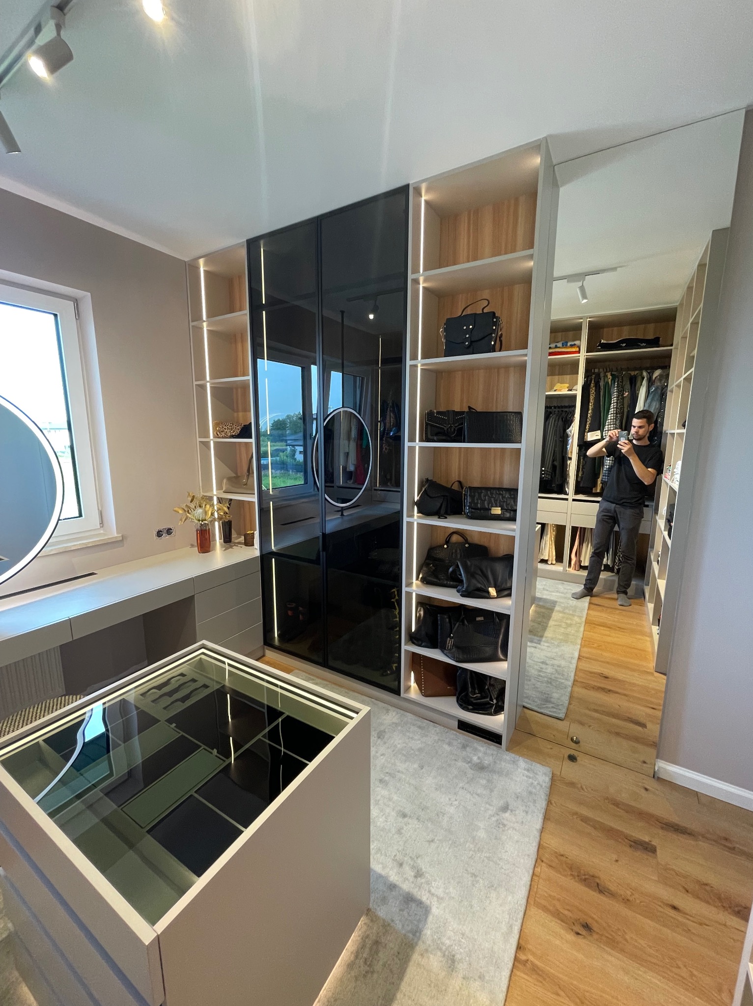 Nowoczesna garderoba na wymiar z czarnymi, szklanymi frontami i oświetleniem LED. Widoczny mężczyzna robiący zdjęcie wnętrza. Na półkach torebki.