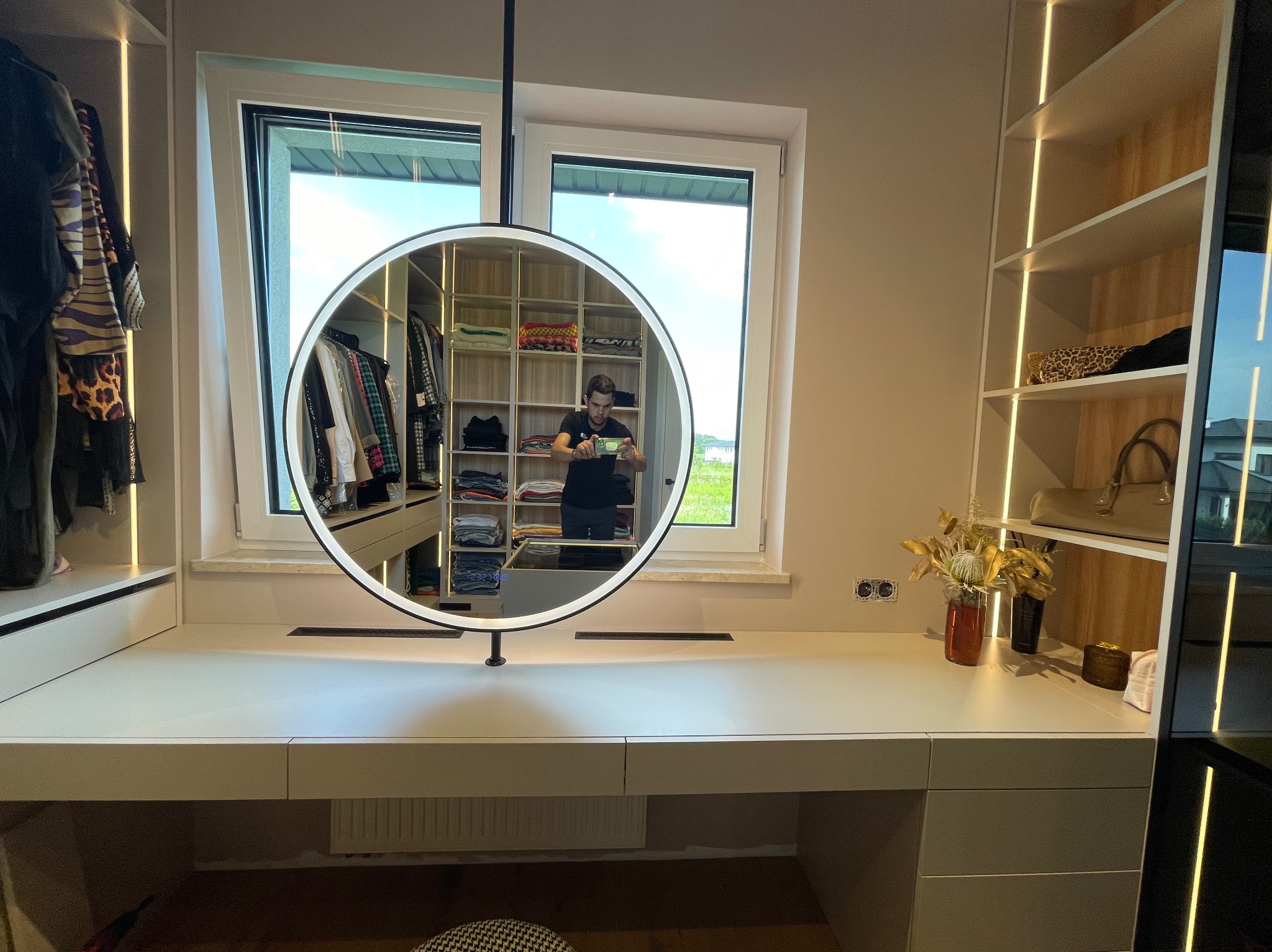 Beżowa garderoba na wymiar z oświetleniem LED, widoczna w okrągłym lustrze. Na blacie dekoracje i gniazdka elektryczne. W tle widok z okna na zieleń.