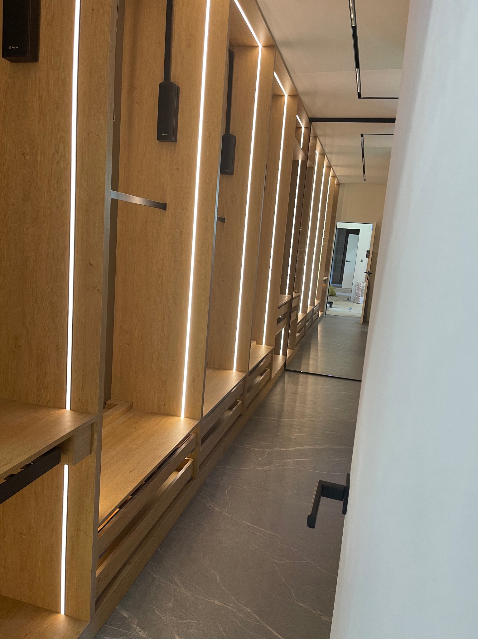 Garderoba na wymiar z oświetleniem LED, drewniane panele, szuflady z uchwytami, szara podłoga. Widok z perspektywy wnętrza, lustro na końcu korytarza.