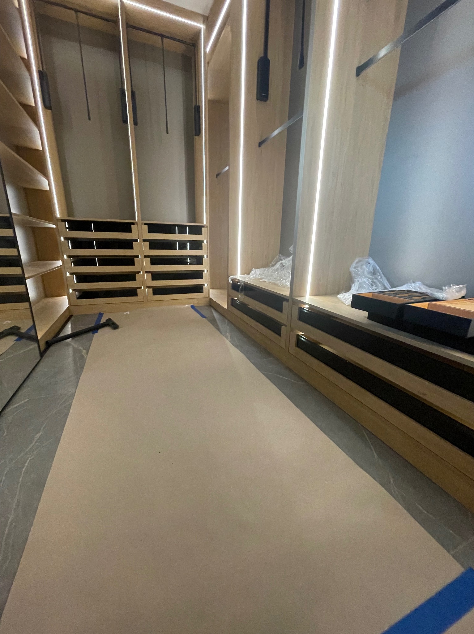 Wnętrze garderoby na wymiar w trakcie montażu, widoczne półki, szuflady z czarnymi uchwytami i oświetlenie LED. Podłoga zabezpieczona kartonem.