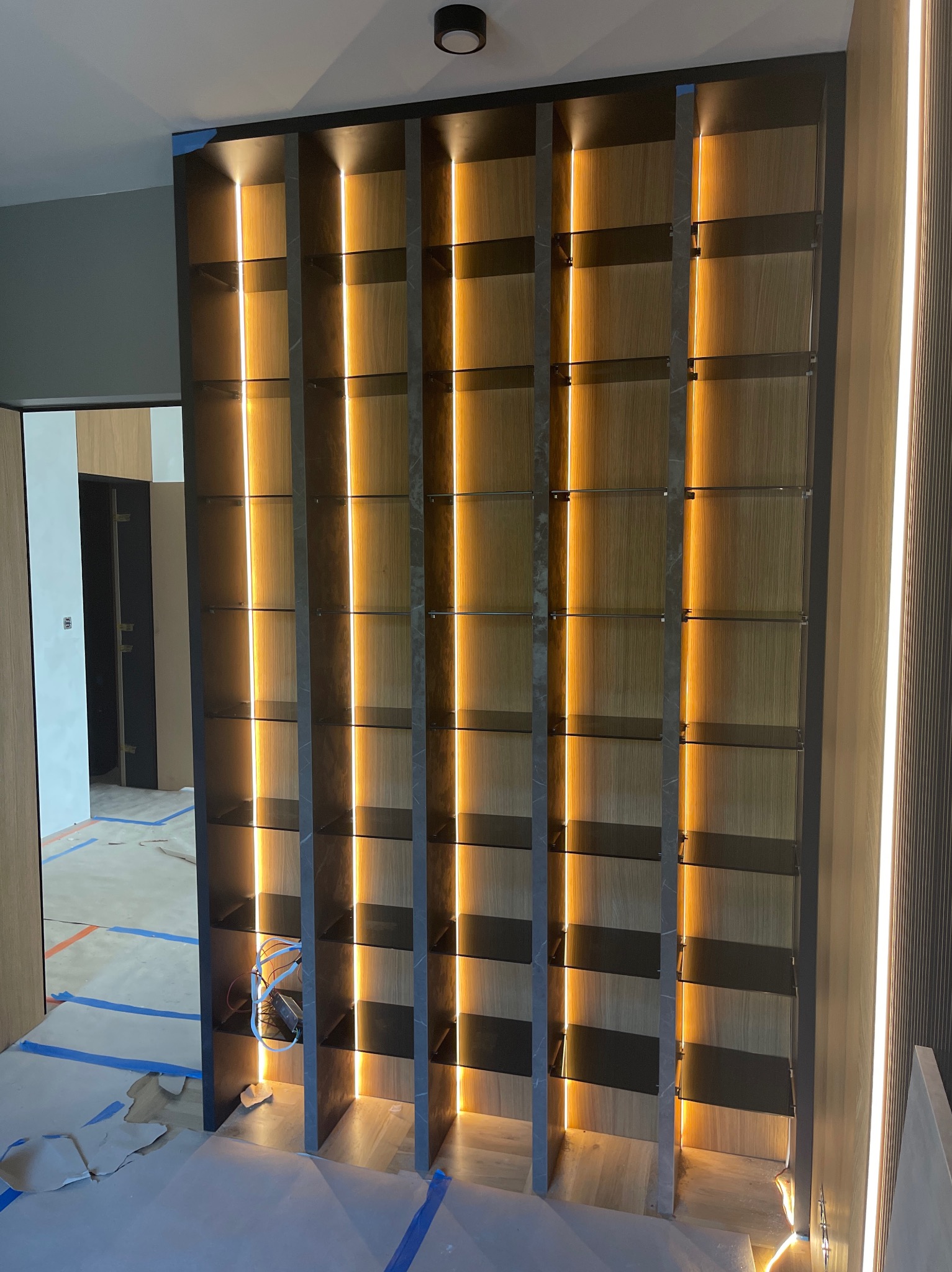 Nowoczesna, podświetlana półka z drewna i szkła w trakcie montażu. Widoczne zabezpieczenia podłogi i kable. Minimalistyczny design, ciepłe oświetlenie.