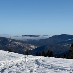 RUDZKI SPÓŁKA Z OGRANICZONĄ ODPOWIEDZIALNOŚCIĄ - Zimowy krajobraz górski z widocznymi śladami na śniegu, lasem iglastym i mgłą w dolinach, uchwycony w słoneczny dzień z perspektywy pieszego szlaku.