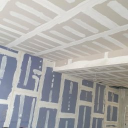 DARBUD Dariusz Baran - Ściana i sufit z płyt gipsowo-kartonowych, przygotowane do szpachlowania. Widoczne łączenia płyt wypełnione masą szpachlową. W tle drabina i przewody.