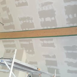 DARBUD Dariusz Baran - Sufit po szpachlowaniu, widoczne łączenia płyt gipsowych. Na pierwszym planie rusztowanie, w tle drabina. Drewniana belka z zieloną taśmą maskującą.