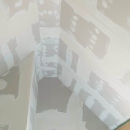 DARBUD Dariusz Baran - Skośny sufit poddasza w trakcie prac wykończeniowych, pokryty warstwą szpachli w jasnych odcieniach szarości i bieli, widoczne drewniane elementy konstrukcyjne.