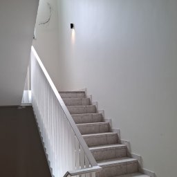 Kameleon Studio - Jasne, odnowione schody z białymi ścianami i poręczą. Minimalistyczne oświetlenie punktowe. Widoczne ślady po instalacji elektrycznej.