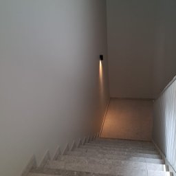 Kameleon Studio - Schody wykończone jasnym marmurem, ściany pomalowane na biało, oświetlenie punktowe na ścianie. Widok z góry na dół, minimalistyczny design.