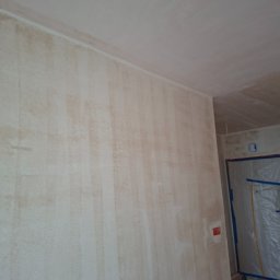 Kameleon Studio - Ściana po szpachlowaniu z widoczną fakturą, przygotowana do dalszych prac wykończeniowych. Widoczny fragment sufitu i zabezpieczone folią drzwi.