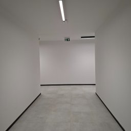 Kameleon Studio - Biały korytarz z szarą podłogą z płytek, oświetlony lampą LED. Widoczny znak ewakuacyjny. Minimalistyczne wykończenie wnętrza, świeżo po remoncie.