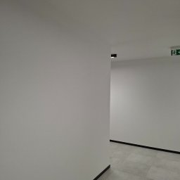 Kameleon Studio - Białe ściany korytarza z szarą podłogą z płytek. Widoczny znak ewakuacyjny i czarne listwy przypodłogowe. Minimalistyczne oświetlenie sufitowe.