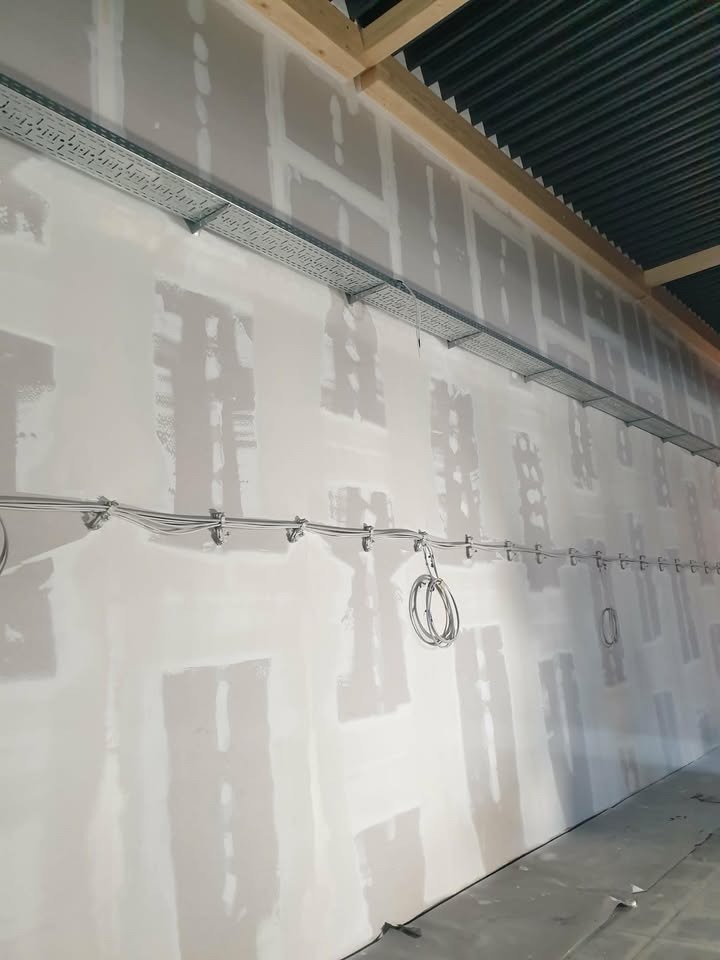 Ściana po szpachlowaniu z widocznymi łączeniami płyt gipsowych, przygotowana do dalszych prac wykończeniowych. Widoczne koryta kablowe i przewody.