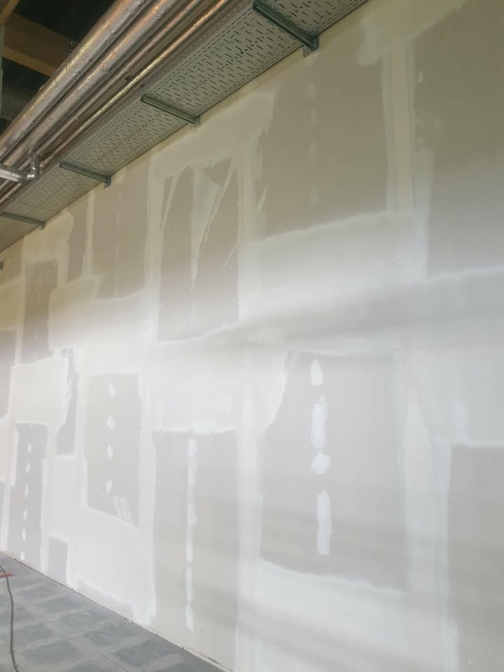 Ściana w trakcie szpachlowania z widocznymi łatami gipsowymi i metalową instalacją pod sufitem. Widok z dołu, surowe wykończenie wnętrza.