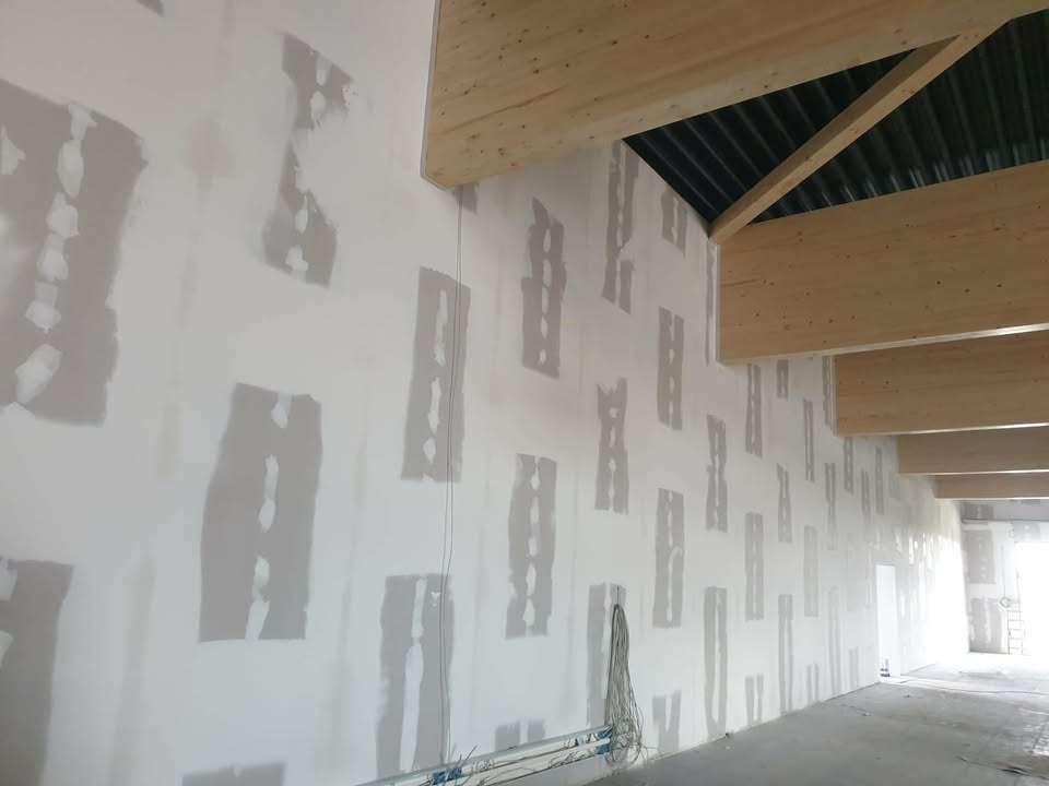 Ściana po szpachlowaniu z widocznymi łatami szpachli, drewniane belki stropowe i kable zwisające ze ściany. W tle widoczne otwarte przestrzenie.