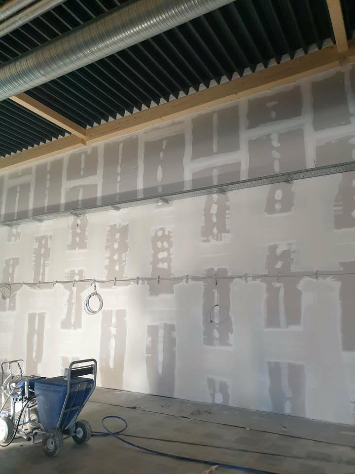 Ściana po szpachlowaniu z widocznymi łatami, przygotowana do dalszych prac wykończeniowych. Na podłodze stoi agregat malarski z niebieskim zbiornikiem.