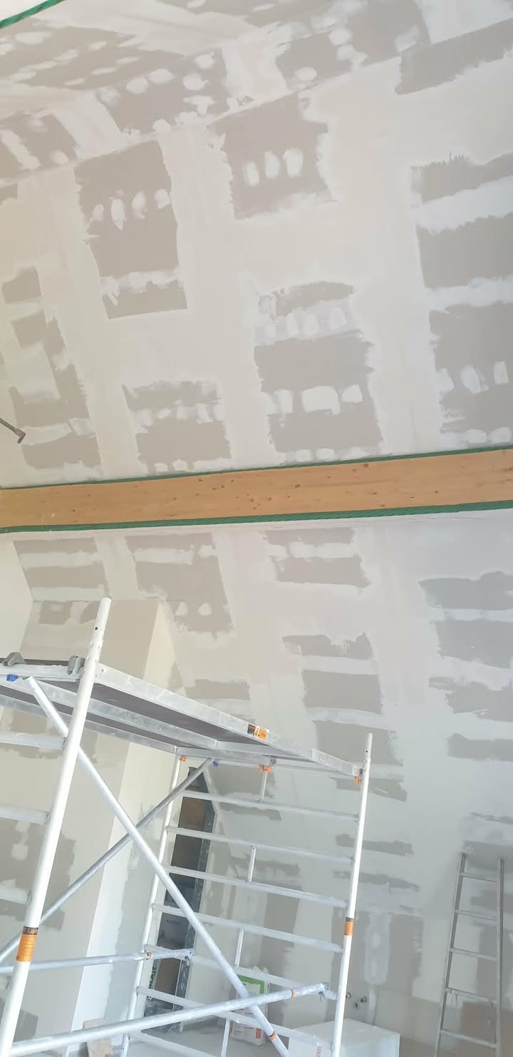 Sufit po szpachlowaniu, widoczne łączenia płyt gipsowych. Na pierwszym planie rusztowanie, w tle drabina. Drewniana belka z zieloną taśmą maskującą.