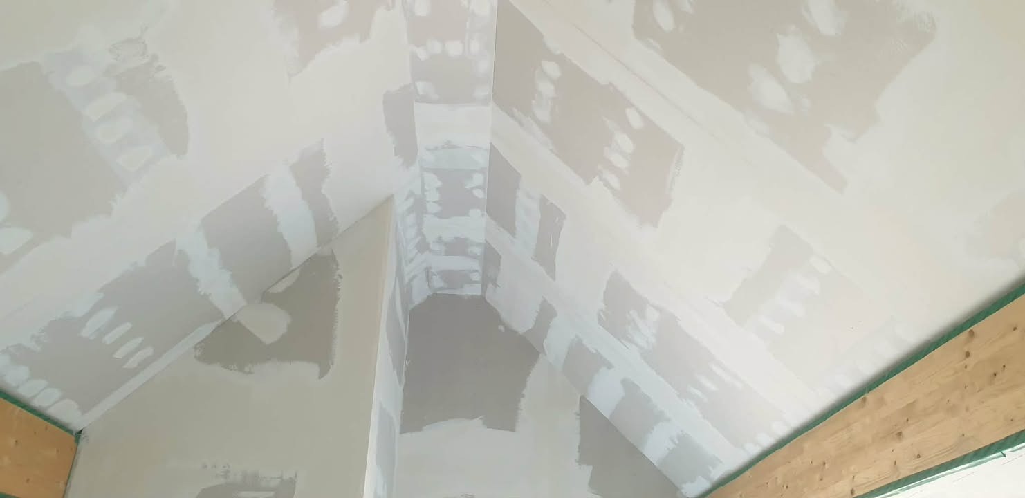 Skośny sufit poddasza w trakcie prac wykończeniowych, pokryty warstwą szpachli w jasnych odcieniach szarości i bieli, widoczne drewniane elementy konstrukcyjne.