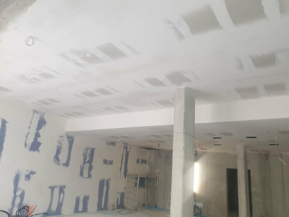 Wnętrze w trakcie prac wykończeniowych: ściany i sufit z widocznymi łatami po szpachlowaniu, przygotowane do dalszych prac. Widoczne rusztowanie i kable.