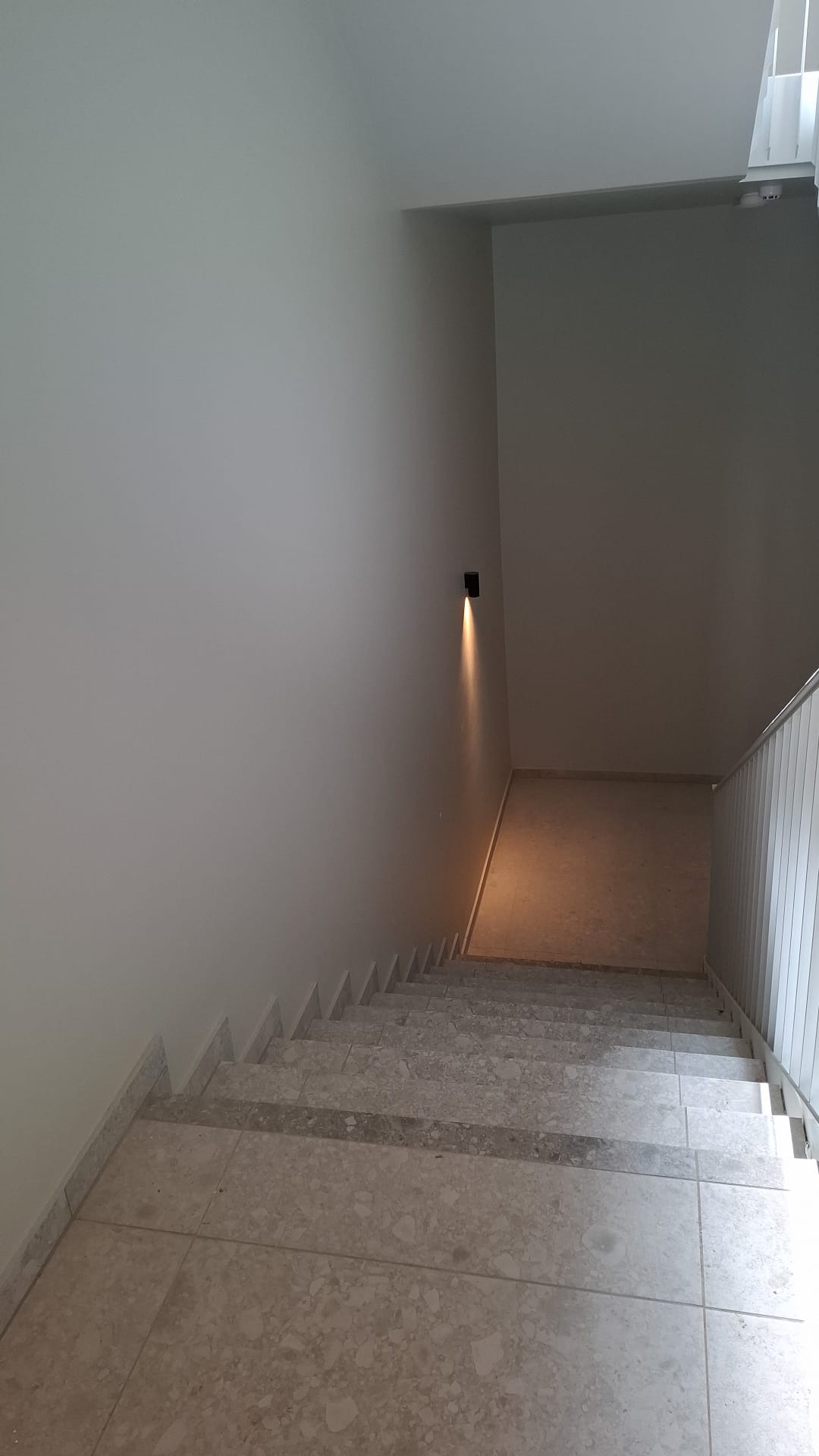 Schody wykończone jasnym marmurem, ściany pomalowane na biało, oświetlenie punktowe na ścianie. Widok z góry na dół, minimalistyczny design.