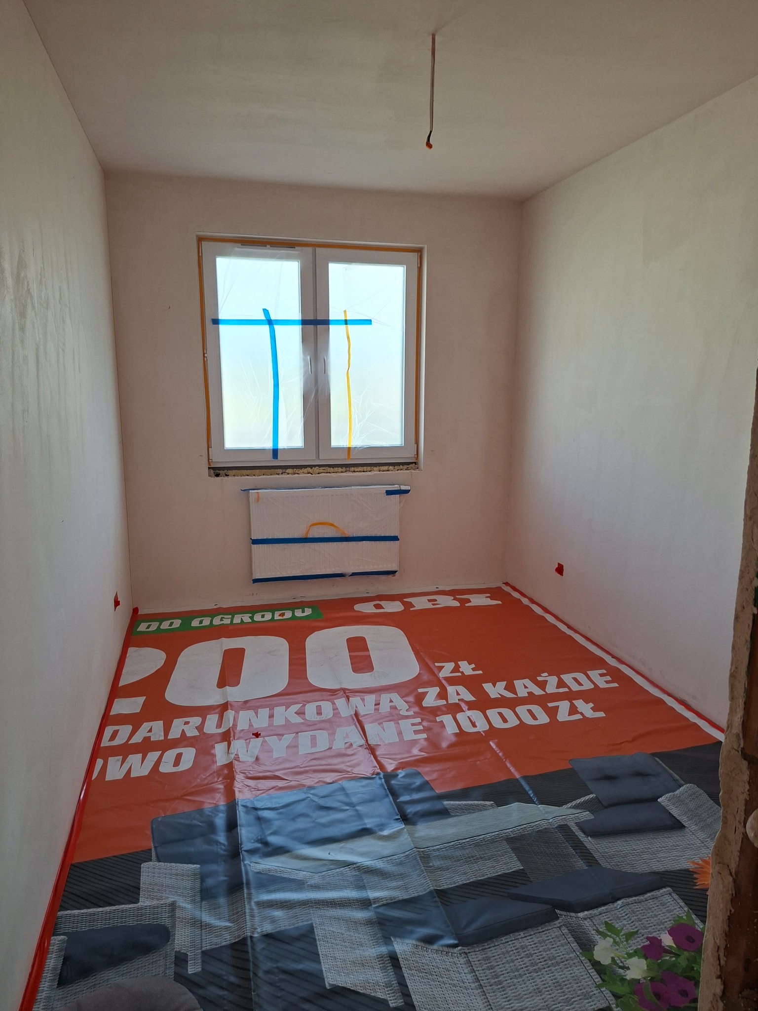 Pokój po szpachlowaniu ścian, zabezpieczony folią ochronną na podłodze i oknach. Widoczne gładkie, jasne ściany przygotowane do malowania. Etap prac wykończeniowych.